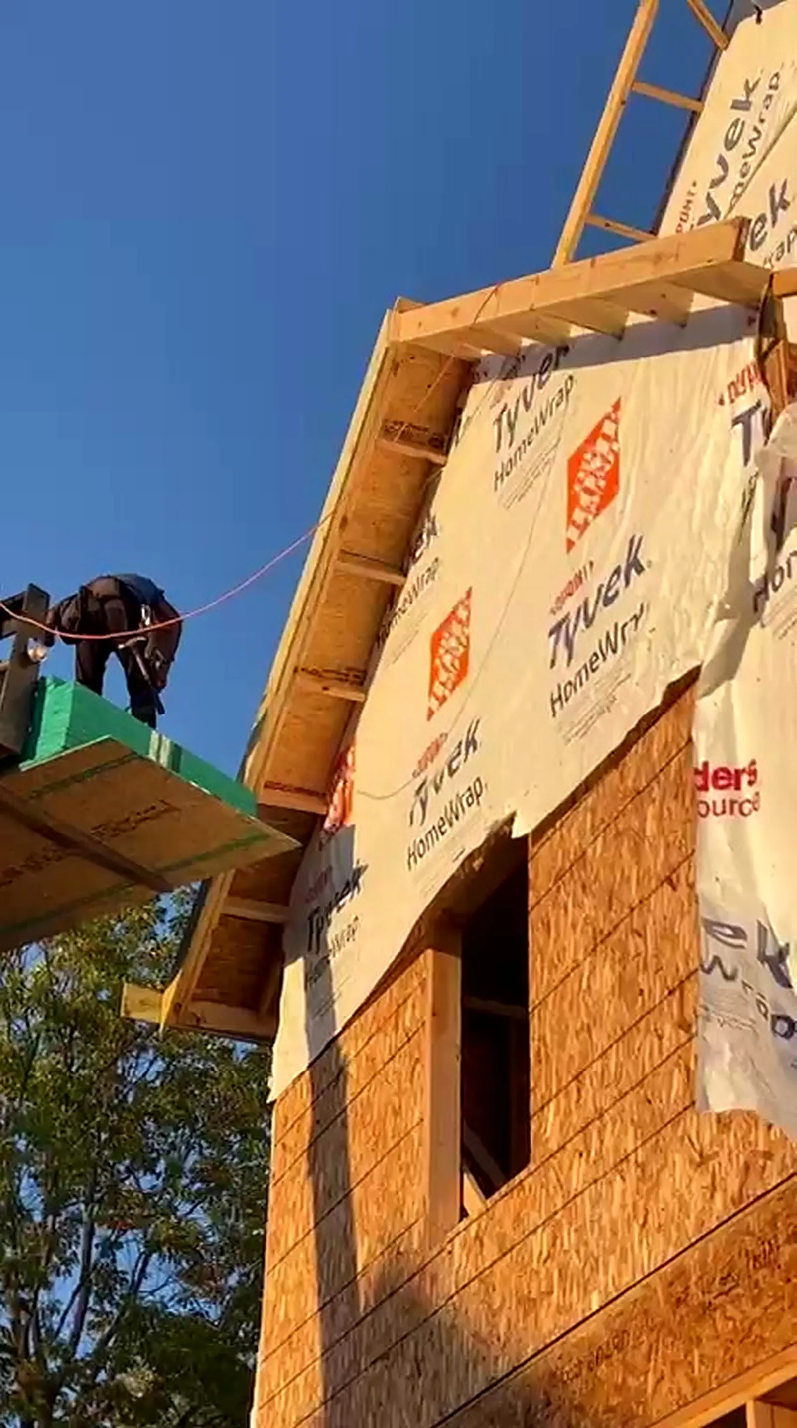 Framer installing house wrap on a gable dormer under a deep blue sky, Berkley MI